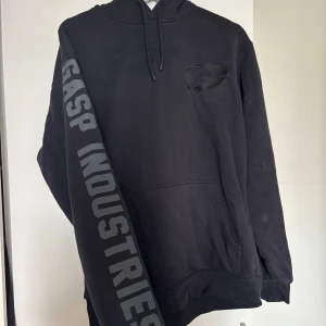 Svart hoodie från GASP Industries - Svart hoodie från GASP Industries med stor logga på ena ärmen och broderad logga på bröstet samt huvan. Klassisk känguruficka framtill, dragsko i huvan och lång ärm. Perfekt för dig som gillar streetwear och sportig stil.
