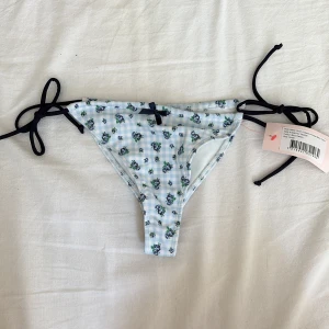 Blommiga bikiniunderdel från Nelly - Supersöt bikiniunderdel från Nelly med ljusblått rutmönster och små blå blommor. Sidorna har svarta knytband och framtill sitter en liten rosett. Perfekt för stranden och sommarens alla bad.