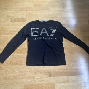 Svart långärmad tröja EA7 Armani - Svart långärmad tröja från EA7 Emporio Armani med stor logga framtill i silver. Tröjan har en klassisk passform och är tillverkad i mjuk bomull, perfekt för en clean och stilren look.