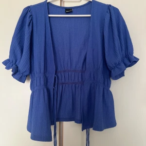 Blå blus med puffärm från Gina Tricot - Supersöt blå blus från Gina Tricot med puffiga korta ärmar och volangdetaljer vid ärmslut och nederkant. Blusen har knytband i midjan och en lätt strukturerad yta som ger extra känsla. Perfekt för dig som vill ha en trendig och luftig look.