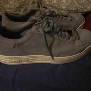 Grå mockasneakers från Adidas - Snygga grå sneakers från Adidas i mocka med klassisk låg profil och vit sula. Skorna har rund tå, snörning och diskret Adidas-logga på sidan av sulan. Perfekta för dig som gillar stilrena och bekväma sneakers med en clean look.