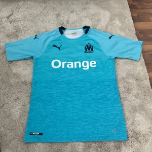 Olympique Marseille  fotbollströja S - Säljer en ljusblå Olympique Marseille fotbollströja från Puma med klubbmärke och sponsortryck på bröstet. Tröjan har korta ärmar, mörkblå detaljer vid kragen och är tillverkad i ett lätt, ventilerande funktionsmaterial. Perfekt för dig som älskar fransk fotboll.
