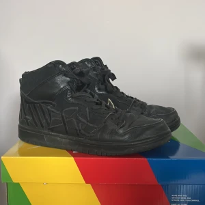 Nike Dunk High SB x Run The Jewels - Unika Nike Dunk High SB sneakers i helsvart skinn med guldiga detaljer och broderad text på hälen. Skorna har snörning, högt skaft och coola grafiska inslag på sidorna. Perfekta för dig som vill sticka ut med streetwear och exklusiv design.