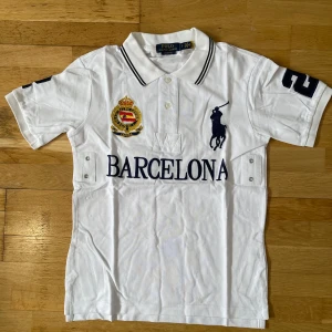 Vit Polo Ralph Lauren Barcelona t-shirt - Vit piké-t-shirt från Polo Ralph Lauren med broderad logga, stort tryck 'BARCELONA' och emblem på bröstet. Klassisk krage med knappar, svarta detaljer och siffran 2 på ärm och rygg. Tillverkad i bomull och har en sportig vibe. passar storlek m