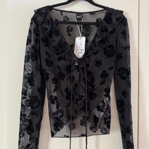 Svart blommig meshblus från Gina Tricot - Supersnygg svart meshblus från Gina Tricot med sammetsrosor, volangkant och snörning framtill.🖤
