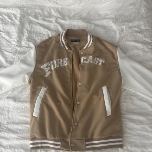 Beige och vit bomberjacka Forecast - Snygg beige och vit bomberjacka från Forecast med broderad text på bröstet. Jackan har vita ärmar, ribbade muddar med ränder och tryckknappar framtill. Perfekt för dig som gillar en sportig och trendig stil.