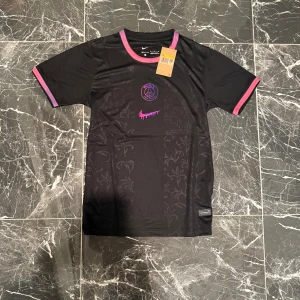 PSG x Nike svart fotbollströja - Svart PSG fotbollströja från Nike med lila och rosa detaljer på krage och ärmslut. Tröjan har ett diskret blommönster i tyget och ett holografiskt PSG-märke samt en rosa Nike-logga med drip-effekt på bröstet. Tillverkad i lätt och ventilerande material.