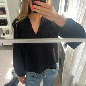 Svart blus med volang från Kappahl - Svart blus från Kappahl med v-ringning, långa puffiga ärmar och snygga volangdetaljer framtill. Blusen har knytband vid halsen,nästintill aldrig använd och i fint skick🥰