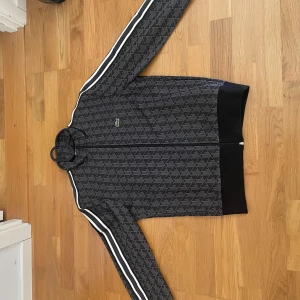 Svart Lacoste tröja  - Köpt från Lacoste hemsida för 2300 Kr. Snygg svart tröja från Lacoste med diskret geometriskt mönster och vita ränder längs ärmarna. Tröjan har dragkedja framtill, ribbade muddar och klassisk krokodillogga på bröstet. Perfekt för dig som gillar sportig och stilren look. 