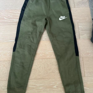 Olivgröna mjukisbyxor från Nike - Snygga olivgröna mjukisbyxor från Nike med svarta paneler längs sidorna och vit Nike-logga framtill. Dock som de syns på bilden finns de två fläckar, något som eventuellt går att ta bort i tvätten. ( vet inte vad det är för fläckar). Annars är resterande mkt fint skick 