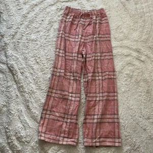 Rosa rutiga pyjamas byxor från H&M - Säljer ett par rosa pyjamas byxor från H&M med rutigt mönster och breda ben. Byxorna har resår i midjan och är gjorda i mjuk bomull, perfekta för en chill och avslappnad stil.
