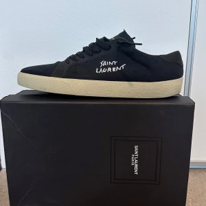 Svarta Saint Laurent sneakers - Svarta sneakers från Saint Laurent med vit broderad logga på sidan och klassisk beige sula. Skorna har snörning och är tillverkade i canvas med detaljer i skinn. Perfekt för dig som gillar stilrena och exklusiva sneakers med streetkänsla. Skorna är i storlek 44 1/2. De har använt max 2-3 gånger 
