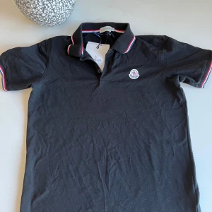 Svart Moncler pikétröja - Stilren pikétröja från lyxmärket Moncler i klassiskt svart med Monclers ikoniska logotyp på bröstet. Trikolor-detaljer (röd, vit & blå) på krage och ärmslut ger en exklusiv och sportig look. Perfekt för både vardag och finare tillfällen.  ✔  verifierbar QR-kod på insidan ✔ Slim fit passform ✔ Material av hög kvalitet – skön och hållbar bomull ✔ Nypris i butik: ca 2 000–2 500 kr ✔️ storlek L passar storlek M 🎁 Skickas snabbt och noggrant paketerad. lyxig tröja till ett mycket bättre pris!  