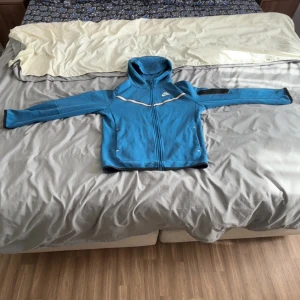 Blå Nike vindjacka med huva 147-158 barn  - Snygg blå vindjacka från Nike med hel dragkedja och huva. Jackan har vita detaljer, två fickor framtill och en extra ficka på ärmen. Perfekt för dig som gillar sportig stil och vill ha något lätt och smidigt.