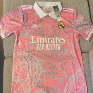 Real Madrid rosa draktröja Adidas - Snygg Real Madrid fotbollströja från Adidas i rosa med grått drakmönster över hela tröjan. Klassisk vit krage, korta ärmar och klubbmärke på bröstet. Tillverkad i lätt och ventilerande material, perfekt för dig som vill sticka ut på planen.