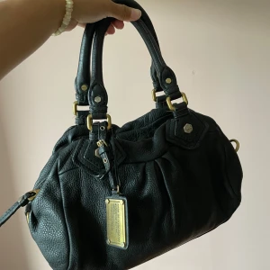 Svart handväska Marc by Marc Jacobs - Säljer en svart handväska från Marc by Marc Jacobs i äkta skinn med snygga guldfärgade metalldetaljer och dubbla handtag. Väskan har en klassisk, rundad form och en metallbricka framtill med märkets logga. Perfekt för dig som gillar stilrena accessoarer.