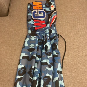BAPE blå camo vindjacka shark hoodie - Säljer en ikonisk BAPE vindjacka i blå camo med hajmotiv på huvan och broderade bokstäver. Jackan har dragkedja framtill, coola detaljer och är perfekt för dig som vill sticka ut. Materialet är syntet och jackan har en loose passform.