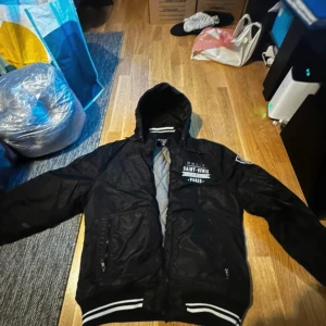 Svart bomberjacka Saint-Denis Paris - Svart bomberjacka med huva och vita detaljer från Saint-Denis Paris. Jackan har tryck på bröstet och ärmen, quiltat grått foder och dragkedjor på fickorna. Ribbstickade muddar med vita ränder ger en sportig vibe. Perfekt för dig som gillar streetstyle.