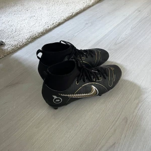  Nike Mercurial Superfly  -  Nike Mercurial Superfly  med guld- och vitdetaljer samt klassisk Swoosh på sidan. Skorna har snörning, högre skaft och mönstrad ovandel i syntetmaterial. Perfekta för dig som vill sticka ut på planen med stilrena och sportiga skor. 