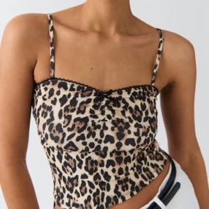 Leopardmönstrad linnetopp med rosett - Säljer en leopardmönstrad linnetopp med tunna axelband, liten svart rosett och spetskant upptill. Toppen är figurnära och har en lätt glansig finish som ger en trendig vibe. Perfekt att styla med jeans eller kjol för en cool look.