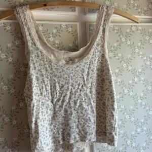 Blommig spetskantad linnetopp Brandy Melville - Supersöt linnetopp från Brandy Melville i vit med små blå blommor. Toppen har spetskant runt halsringning och axelband, vilket ger en romantisk vibe. Tillverkad i mjuk bomull, perfekt för varma dagar eller lager på lager.