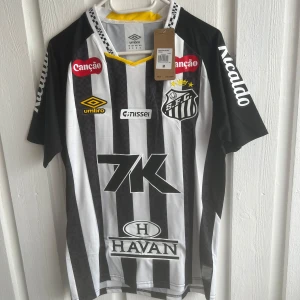 Santos FC Neymar Jr matchtröja Umbro - Santos FC fotbollströja från Umbro med Neymar Jr och nummer 10 på ryggen. Klassisk svartvit randig design med gula detaljer och flera sponsorloggor. Kortärmad modell i lätt och ventilerande material, perfekt för dig som älskar brasiliansk fotboll.