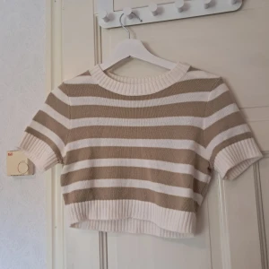 Randig croppad topp från H&M  - Superfin croppad stickad topp från H&M i beige och vitt med breda ränder. Toppen har korta ärmar, ribbade muddar och rund halsringning. Perfekt för dig som gillar en chill och trendig stil. Använd typ en gång