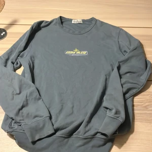Grå Stone Island sweatshirt - Säljer en grå långärmad sweatshirt från Stone Island med broderad logga i gult och vitt på bröstet. Tröjan har rund halsringning och ribbade muddar vid ärmslut och nederkant. Perfekt för dig som gillar streetwear och vill ha en clean look.