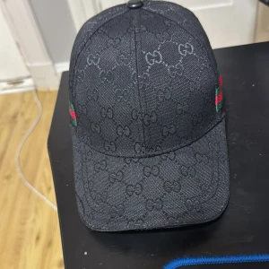 Svart Gucci keps med GG-mönster - Snygg svart keps från Gucci med klassiskt GG-monogram över hela kepsen och gröna/röda ränder på sidorna. Justerbar rem baktill för perfekt passform. Tillverkad i bomull och polyester, perfekt för dig som vill sticka ut med en lyxig accessoar.