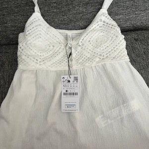 Vit sommartopp från Stradivarius - Supersöt vit topp från Stradivarius med virkade detaljer upptill, smala axelband och knappar framtill. Perfekt för varma dagar och en fräsch look.