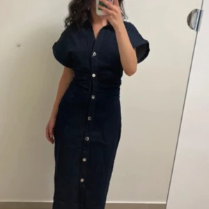 Mörkblå jeansklänning med knappar S - Snygg mörkblå jeansklänning i storlek S. Klänningen har korta ärmar, markerad midja och knappar hela vägen framtill. Klassisk skjortkrage och rak, lång siluett som ger en cool och avslappnad look.