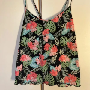 Blommig linnetopp från H&M - Snygg linnetopp från H&M med tropiskt blommönster i rosa, grönt och turkost på svart botten. Tunt tyg i bomull och viskos, smala axelband och lätt v-ringad hals. Perfekt för varma dagar och sommarvibbar.