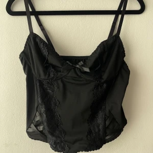 Svart korsett-topp med spetsdetaljer - Snygg svart korsett-topp med tunna justerbara axelband, spetsdetaljer framtill och liten rosett vid bysten. Sidorna har dekorativa band och tyget är glansigt med en croppad passform. 
