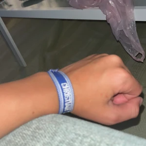 Blått christian dior armband - Snyggt blått tygarmband från Christian Dior med texten 'J'ADIOR' broderad i vitt. Armbandet har justerbar knytning med tofsar i ändarna och en cool, avslappnad vibe. Perfekt accessoar för att lyfta din outfit med en touch av lyx.