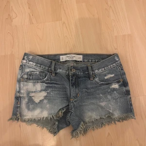 Jeansshorts från Abercrombie & Fitch - Snygga blå jeansshorts från Abercrombie & Fitch med slitna detaljer, fransig kant och klassisk femficksdesign. Shortsen har låg midja och är tillverkade i denim med coola slitningar och ljusa partier för en avslappnad vibe.