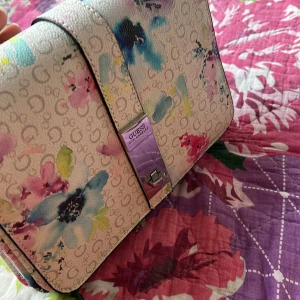 Blommig axelväska från Guess - Superfin axelväska från Guess med blommigt mönster. Väskan är endast använd ett antal par gånger. Perfekt storlek för det viktigaste och ger en färgklick till din outfit.