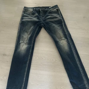 Replay jeans i mörkblå tvätt - Snygga Replay jeans i mörkblå tvätt med slitningar och lagom distressed look på benen. Klassisk femficksmodell med raka ben och tydliga kontrastsömmar. Jeansen har Replay-logga på fickan och bakfickor med dekorativa sömmar. Perfekta för en avslappnad streetstil. 