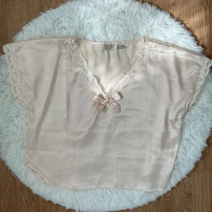 Champagnefärgad blus från Victoria's Secret (vintage) - Söt champagnefärgad blus från Vintage Victoria's Secret i glansig polyester med spetsdetaljer längs v-ringningen och ärmarna. Luftig och ärmlös modell som känns lyxig och feminin. Storlek L(40-42) men passar även storlek M(38) om du vill ha en oversize look.