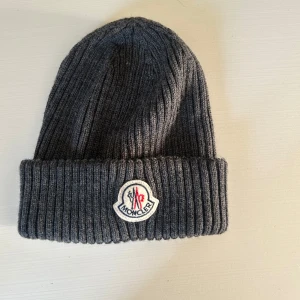 Mörkgrå ribbstickad Moncler mössa - Snygg mörkgrå ribbstickad mössa från Moncler med klassisk uppvikt kant och broderad logotyp framtill. Perfekt för kalla dagar och ger en clean streetstyle-vibe. Materialet är mjukt och värmande, och passformen är normal.