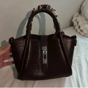 Brun handväska med scarf-detalj - Säljer en snygg mörkbrun handväska som kommer med en dekorativt mönstrat scarf som knyts runt handtaget. Det tillkommer dessutom ett långt band till!😊