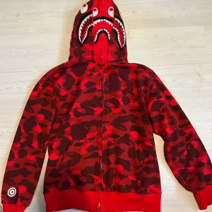 BAPE double zip up röd XL - Ny, XL, ingen skada. Säljer en ikonisk röd camo hoodie från BAPE med hajmotiv på huvan och hel dragkedja. Jackan har unikt camouflagemönster i olika röda nyanser och detaljer som broderade ögon och tänder på huvan. Perfekt statement-plagg för streetwear-entusiasten.