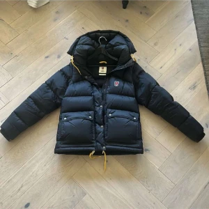 Svart dunjacka Fjällräven Expedition S - Svart dunjacka från Fjällräven Expedition Series med ikonisk röd logga på bröstet. Jackan har huva med gula snören, stora fickor med knappar och dragkedja framtill. Perfekt för kalla dagar och riktigt snygg streetstil.