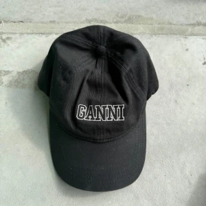 Svart Ganni keps med broderad logga - Svart keps från Ganni i bomull med klassisk böjd skärm och justerbar rem baktill. Använd, däremot inte på senaste året och har mest legat på hyllan. Därmed mycket kvar att ge och väldigt bra skick. Normalt slitage.