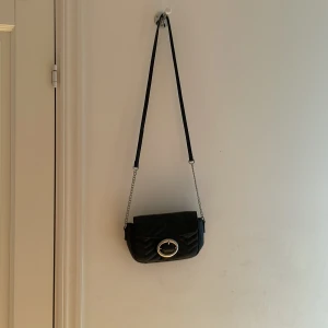 Svart quiltad axelväska med kedja - Snygg svart axelväska i quiltat skinn med silverfärgad kedja och axelrem i läder. Väskan har ett stort spänne framtill i silver och lockstängning. Perfekt storlek för det viktigaste och ger en cool vibe till din outfit.