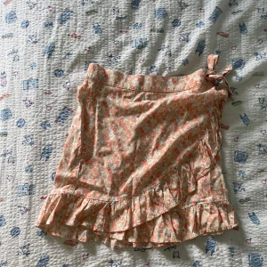 Blommig omlottkjol med volang - Supersöt kort omlottkjol med blommigt mönster i ljusa färger och volangkant. Kjolen har knyt i sidan och är perfekt för varma dagar. Lätt och luftig känsla, passar perfekt till sneakers eller sandaler.