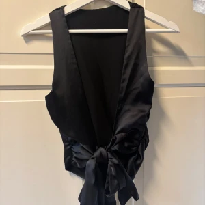 Svart topp med öppen rygg  - Snygg svart topp med öppen rygg i satinliknande material. Långa knytband i bak så man kan knyta själv, man kan även dra runt den ett varv i midjan som ger en cool och trendig look. 