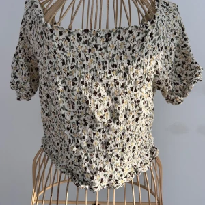 Blommig smockad cropped off shoulder topp från Vero Moda - Supersöt smockad topp från Vero Moda med blommigt mönster i vitt, grönt, gult och brunt. Toppen har korta ärmar, offshoulder-modell och volangkant nertill. Perfekt för dig som gillar romantisk och somrig stil. Passar S och M.