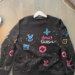 Louis Vuitton svart sweatshirt med färgstänk - Svart sweatshirt från Louis Vuitton med färgglada loggor och stänk i blått, rosa, gult och vitt. Crewneck-modell med lång ärm och klassisk passform. Tryckta LV-symboler och graffiti-inspirerad design gör den riktigt unik. Perfekt för dig som vill sticka ut.