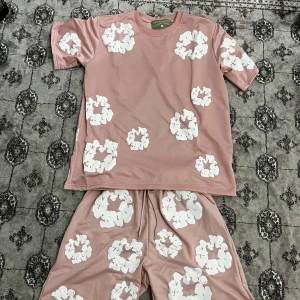   Denim Tears set - Säljer ett par rosa shorts från Denim Tears med stora vita blommiga tryck över hela plagget. Shortsen har snörning i midjan och är gjorda i ett mjukt, lätt material som känns skönt mot huden. Perfekta för en chill och trendig sommarlook.