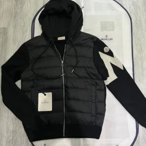 Moncler Cardigan - Säljer en helt ny svart cardigan från Moncler med vadderad framsida, stickade ärmar och huva. Jackan har dragkedja, två fickor fram och en vit detalj på ena ärmen. Perfekt för dig som vill ha en snygg och sportig look. Jackan har NFC på Armen och taggen som man kan scanna.